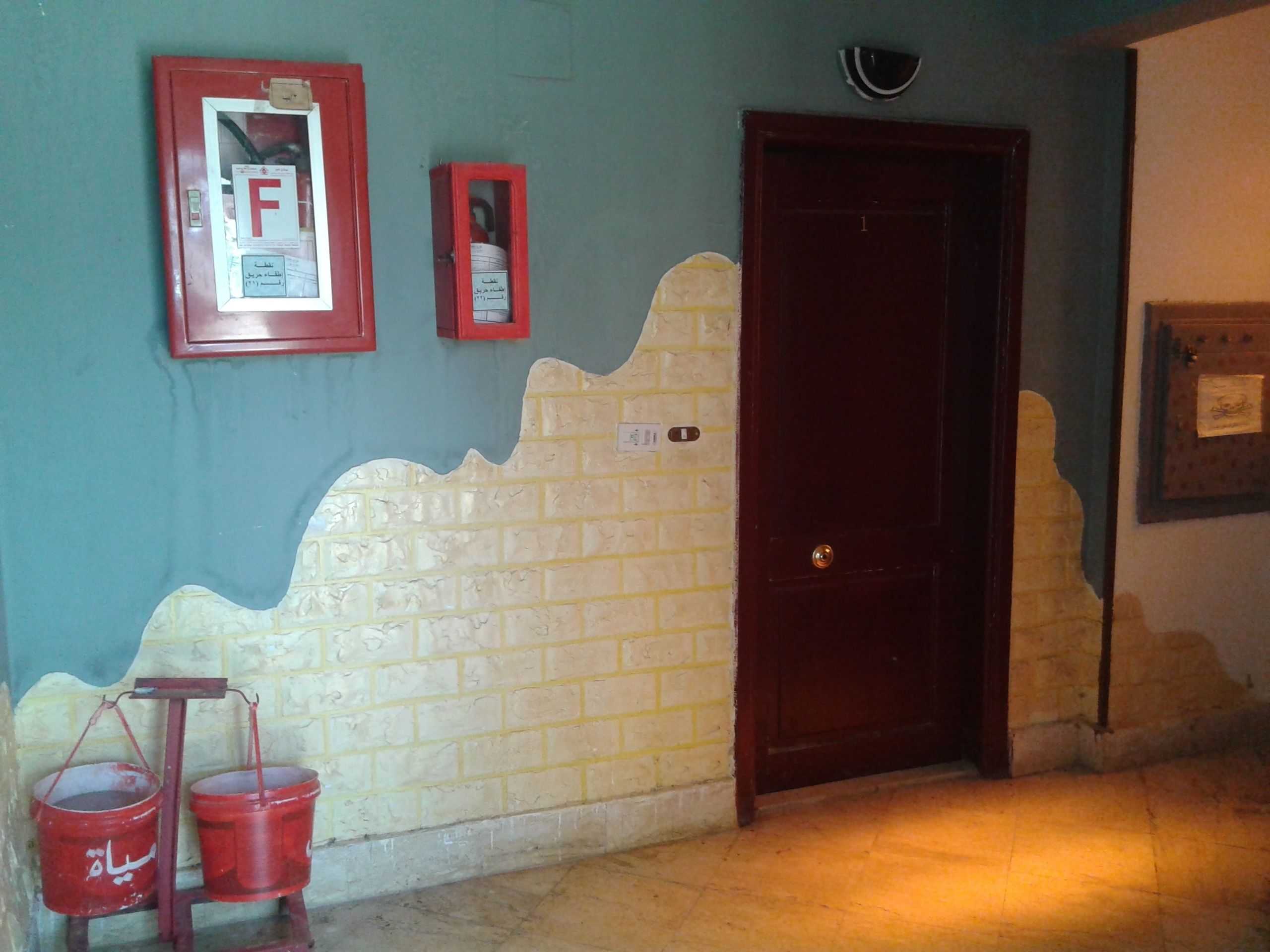 Una habitación con una puerta, un detector de incendios instalado en la pared y dos cubos rojos sobre un soporte.