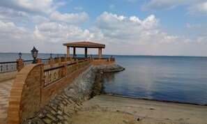 A coastal walkway with a stone railing, a covered pavilion, and a calm sea in the background.