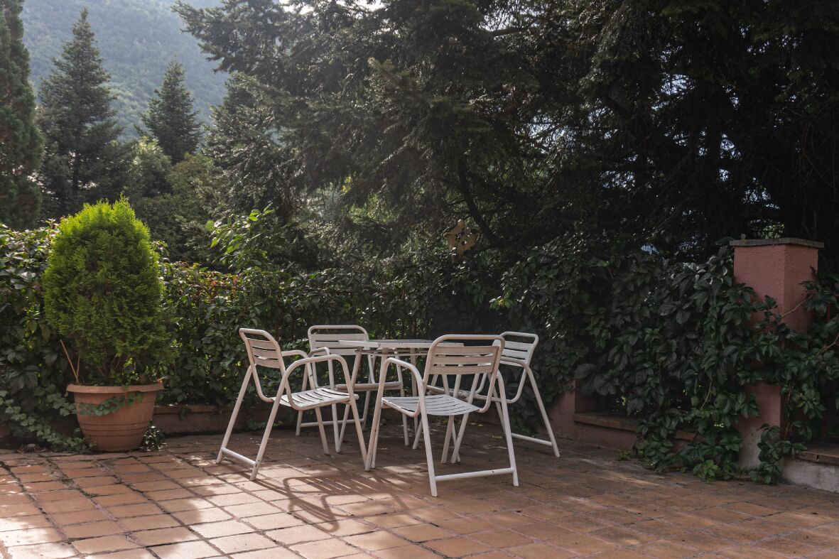 Um pátio externo com uma mesa redonda e quatro cadeiras, rodeado por vasos de plantas e árvores.
