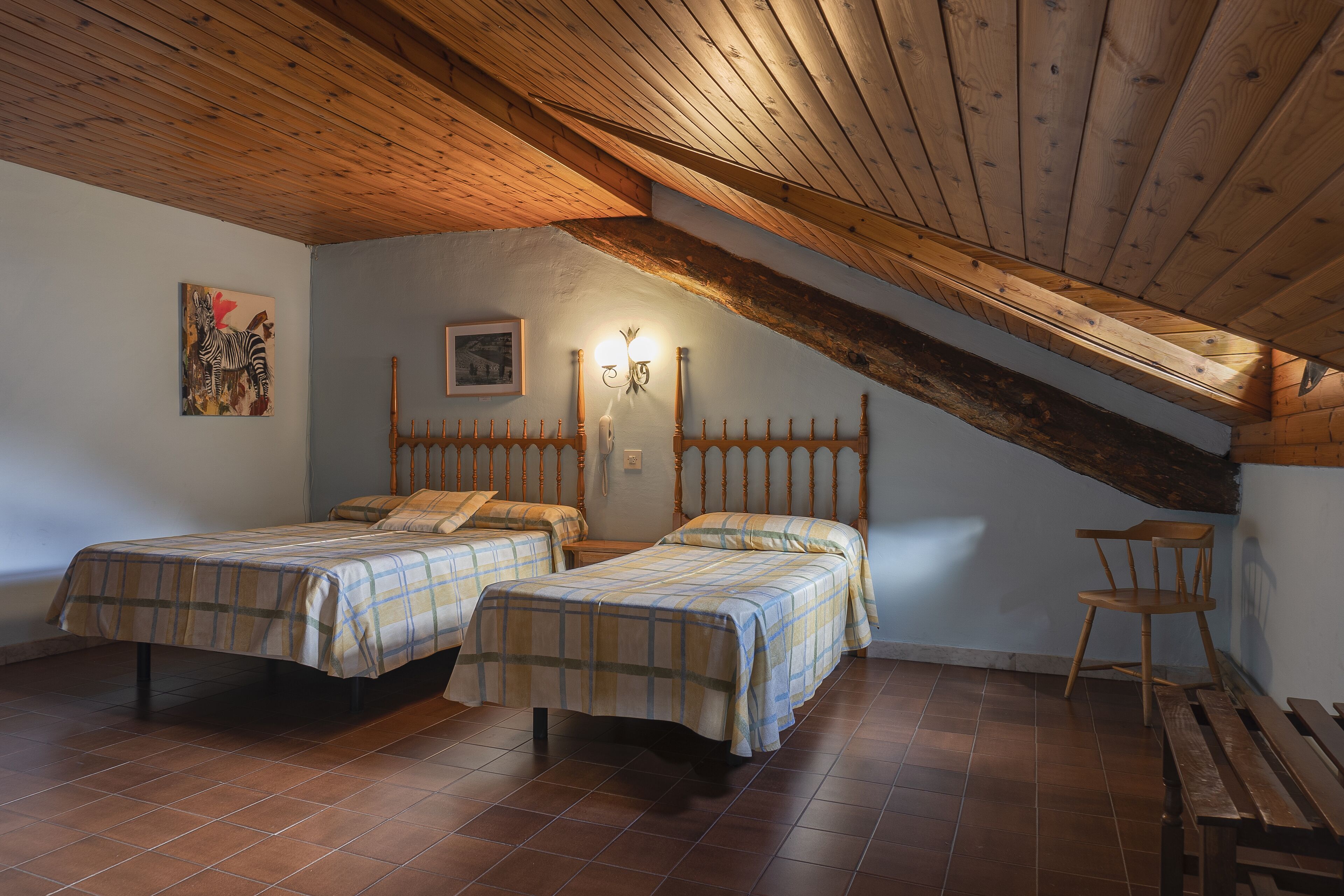 Une chambre avec deux lits, des poutres apparentes, une applique murale et un banc en bois.