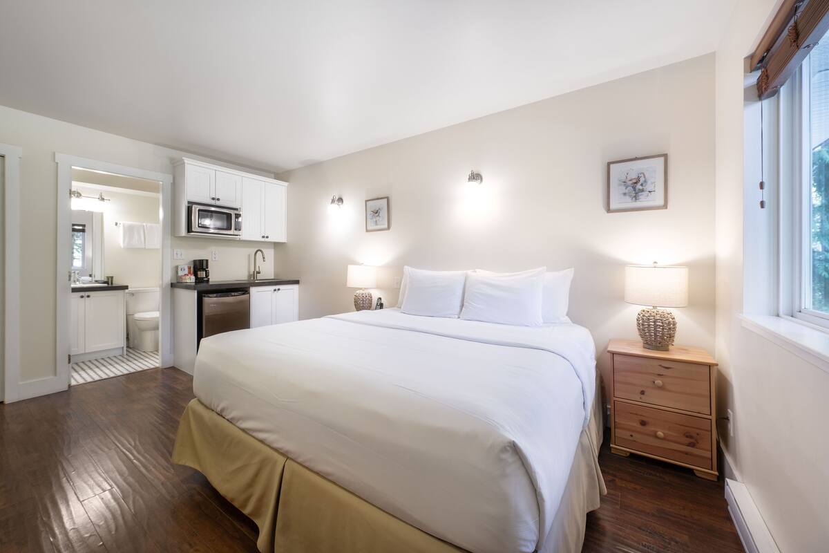 A bedroom with a bed, bedside lamps, a nightstand, and a view of the kitchen.