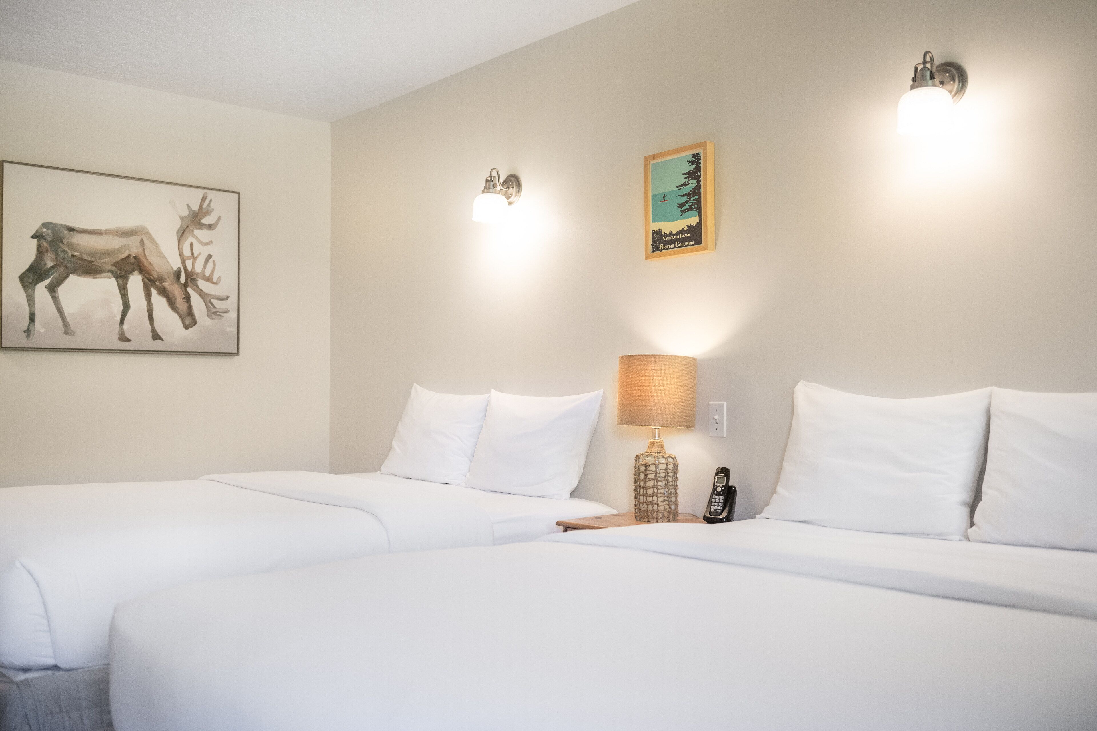 A hotel room with two beds, a wall-mounted lamp, a framed picture of a deer, and a small table with a lamp.