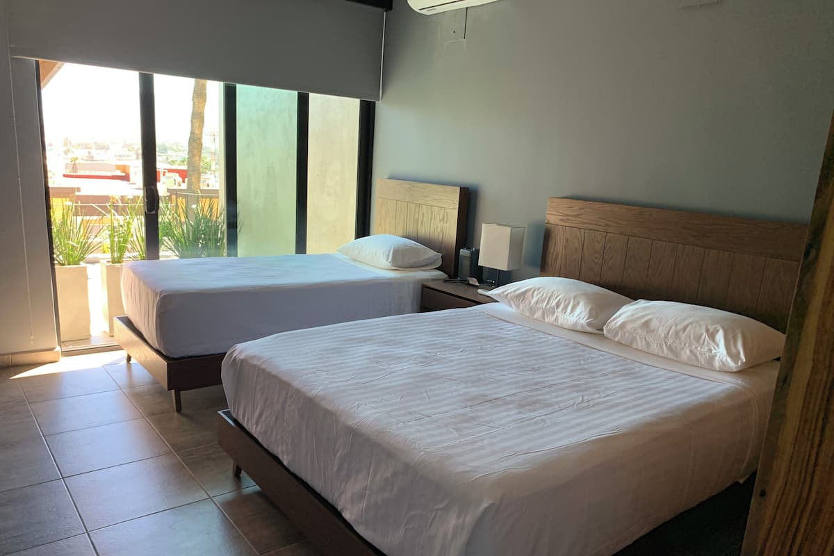 A hotel room with two beds, a nightstand, a lamp, and a view of the outdoors.