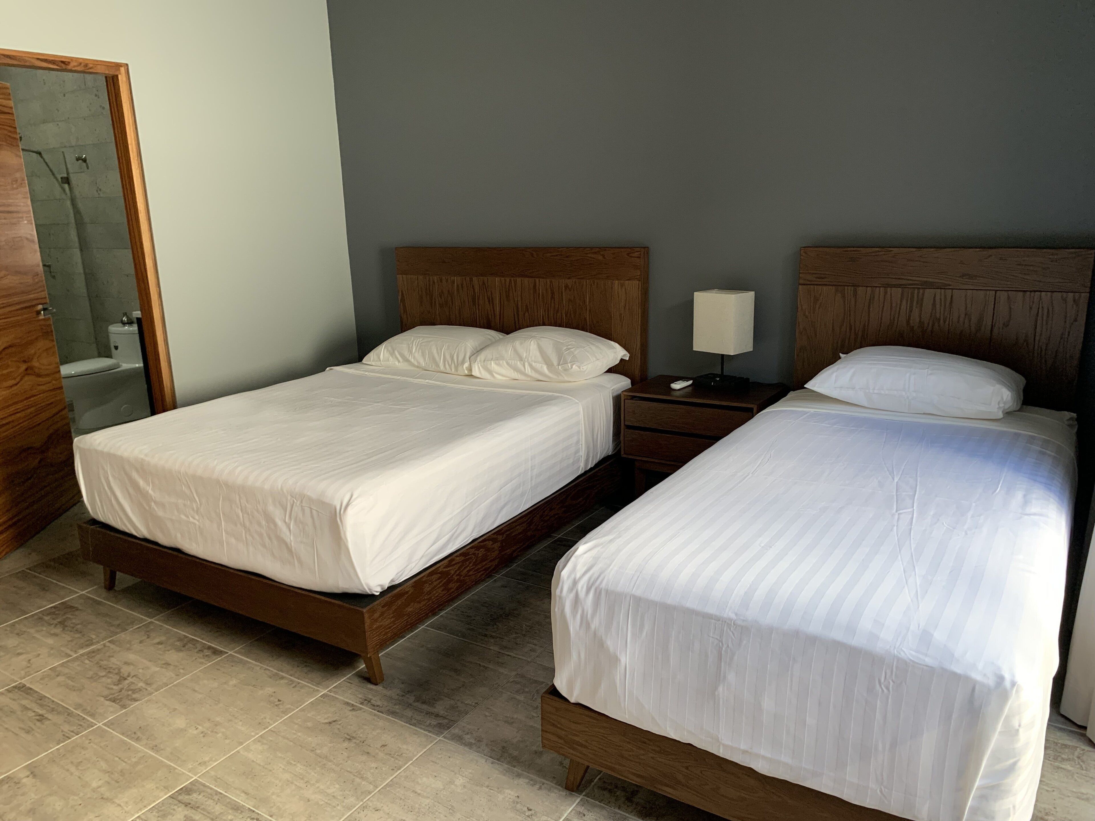 Una cama doble con cabecera de madera, una mesita de noche con lámpara, y un baño visible a través de una puerta abierta.