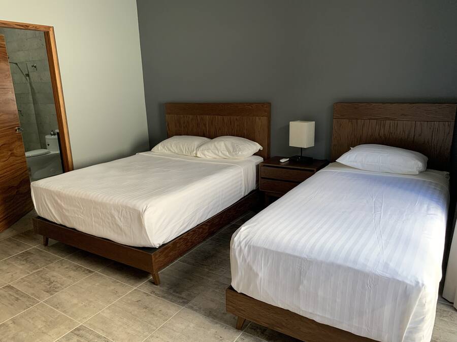 Una cama doble con cabecera de madera, una mesita de noche con lámpara, y un baño visible a través de una puerta abierta.