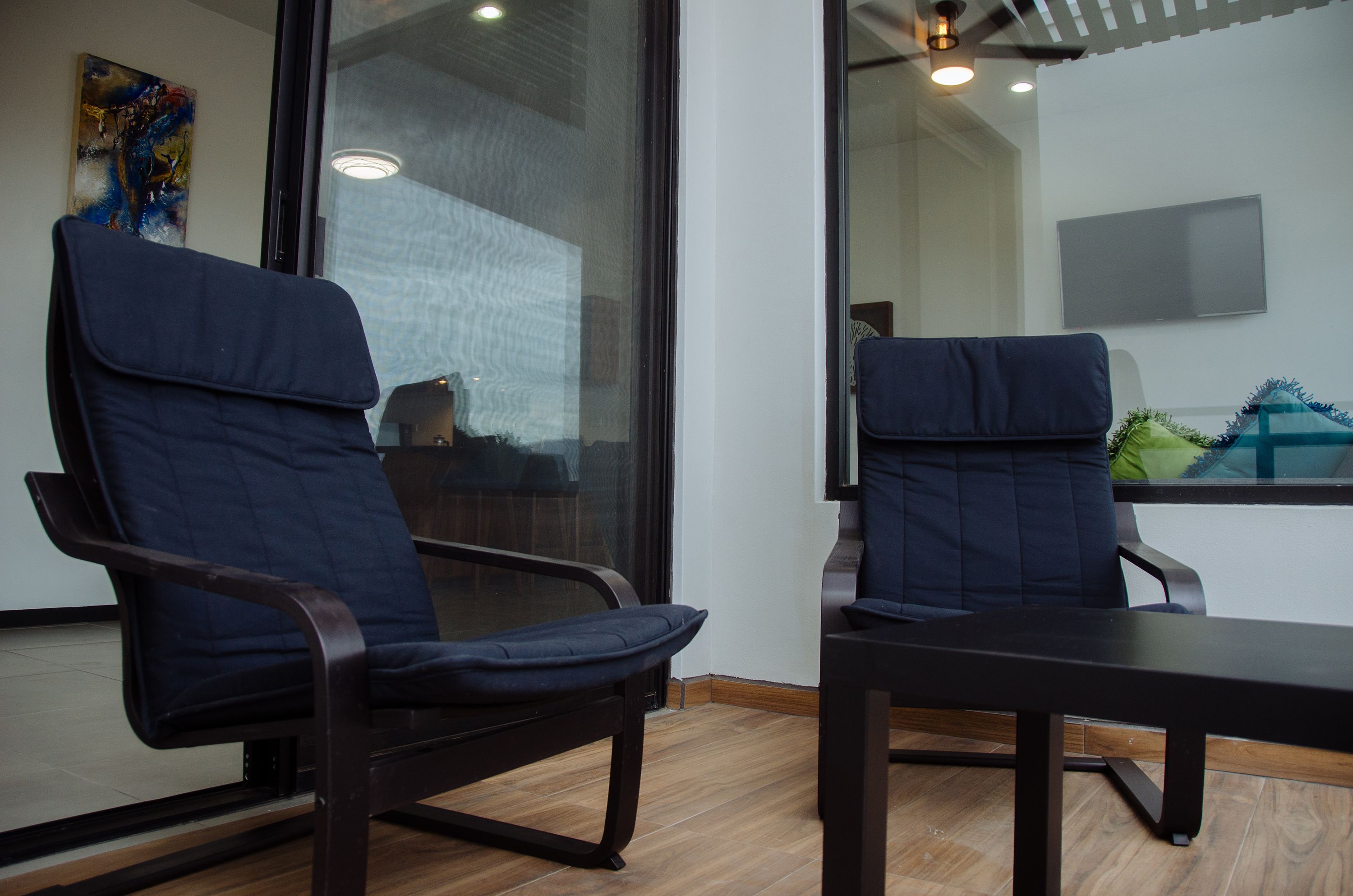 Two dark blue chairs with cushioned seats and backs, a wooden table, and a glass partition with a reflection of a room and a television.