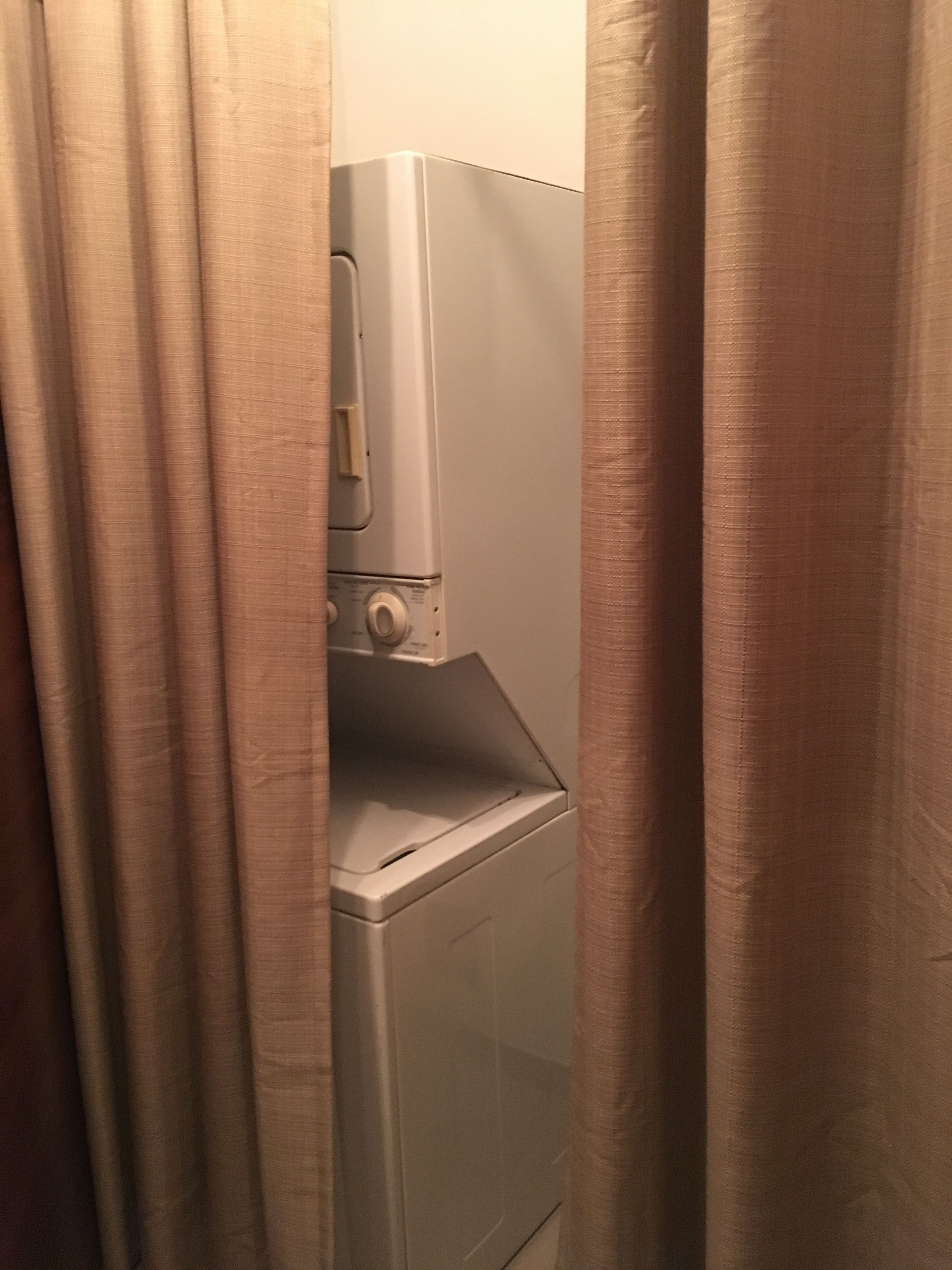 A laundry room with a washing machine and dryer, curtains partially drawn.
