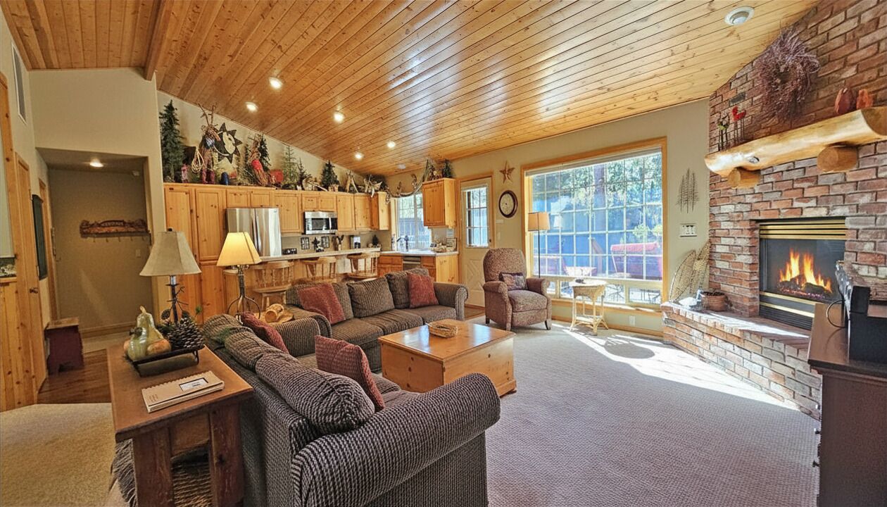 A cozy living room with a fireplace, wooden ceiling, and a kitchen in the background.