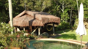 Une construction à toit de chaume, dotée d’une piscine et d’une terrasse en bois, entourée d’une végétation luxuriante.
