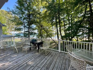 Une terrasse en bois, meublée pour l’extérieur, avec un barbecue et une vue sur les arbres et l’eau.