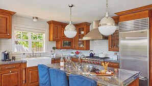 A modern kitchen with wooden cabinets, a stainless steel refrigerator, a large island with bar stools, and a window providing natural light. - Luxurious Boutique Villa w/ Private Pool, Hot Tub, EV Charger in Heart of Delray (Delray Beach)