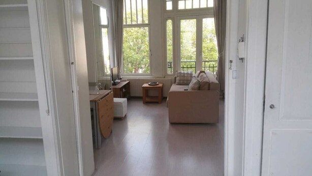 A spacious room with a sofa, a small table, and a window providing a view of greenery outside. - Apartment in Rotterdam (Rotterdam)