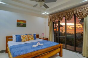 Une chambre à coucher avec un lit, un ventilateur de plafond, un tableau, une table de chevet avec un vase, et une vue sur le coucher de soleil par la fenêtre.