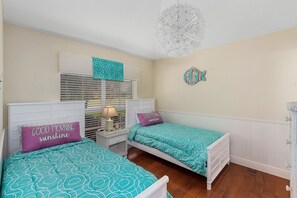 Quarto com duas camas de solteiro, uma mesa de cabeceira com abajur, uma janela com persianas e um painel decorativo na parede.
