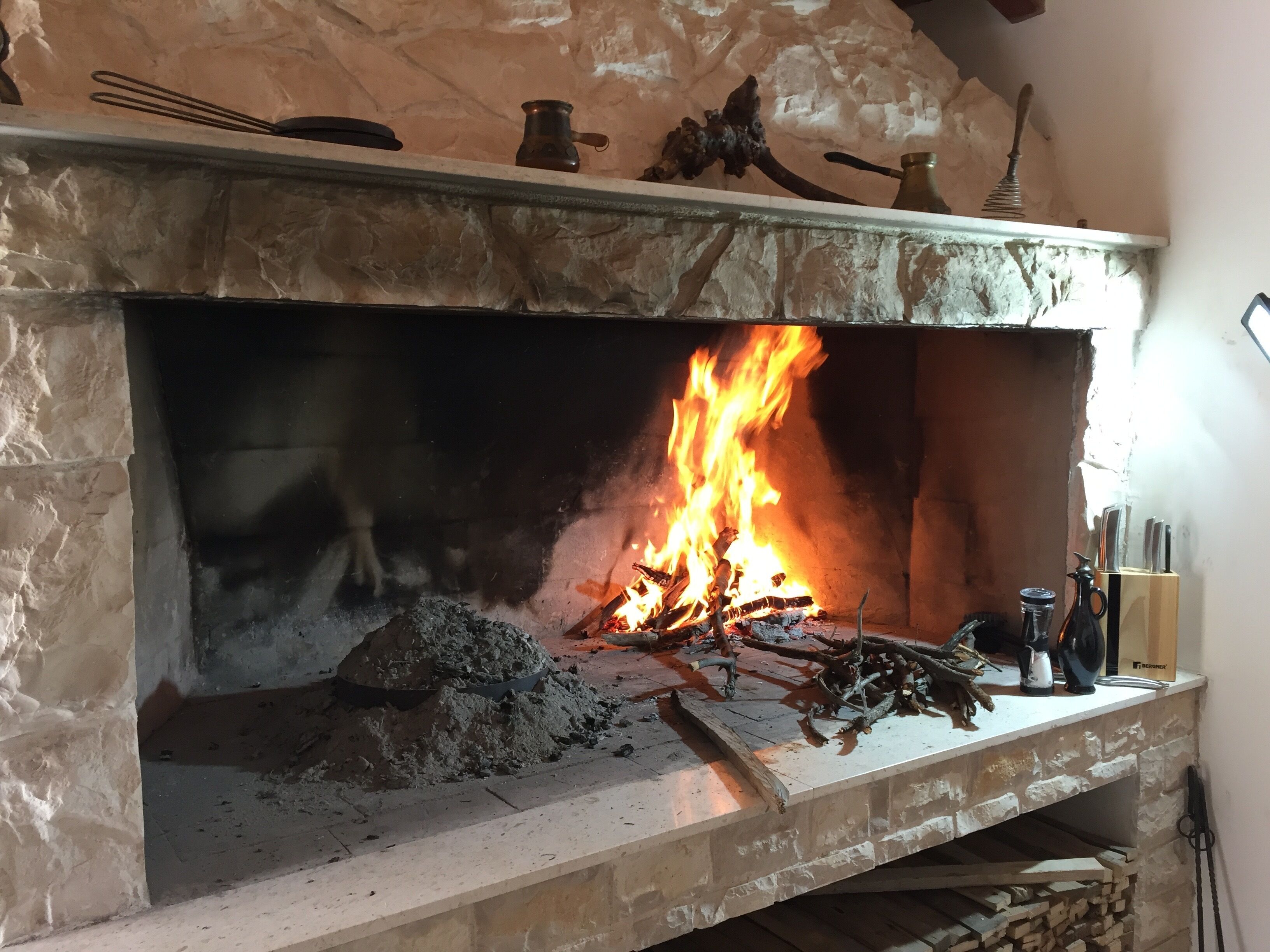Una chimenea de piedra con fuego encendido, rodeada de varios utensilios de cocina y una pila de leña.