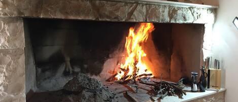 Una chimenea de piedra con fuego encendido, rodeada de varios utensilios de cocina y una pila de leña.