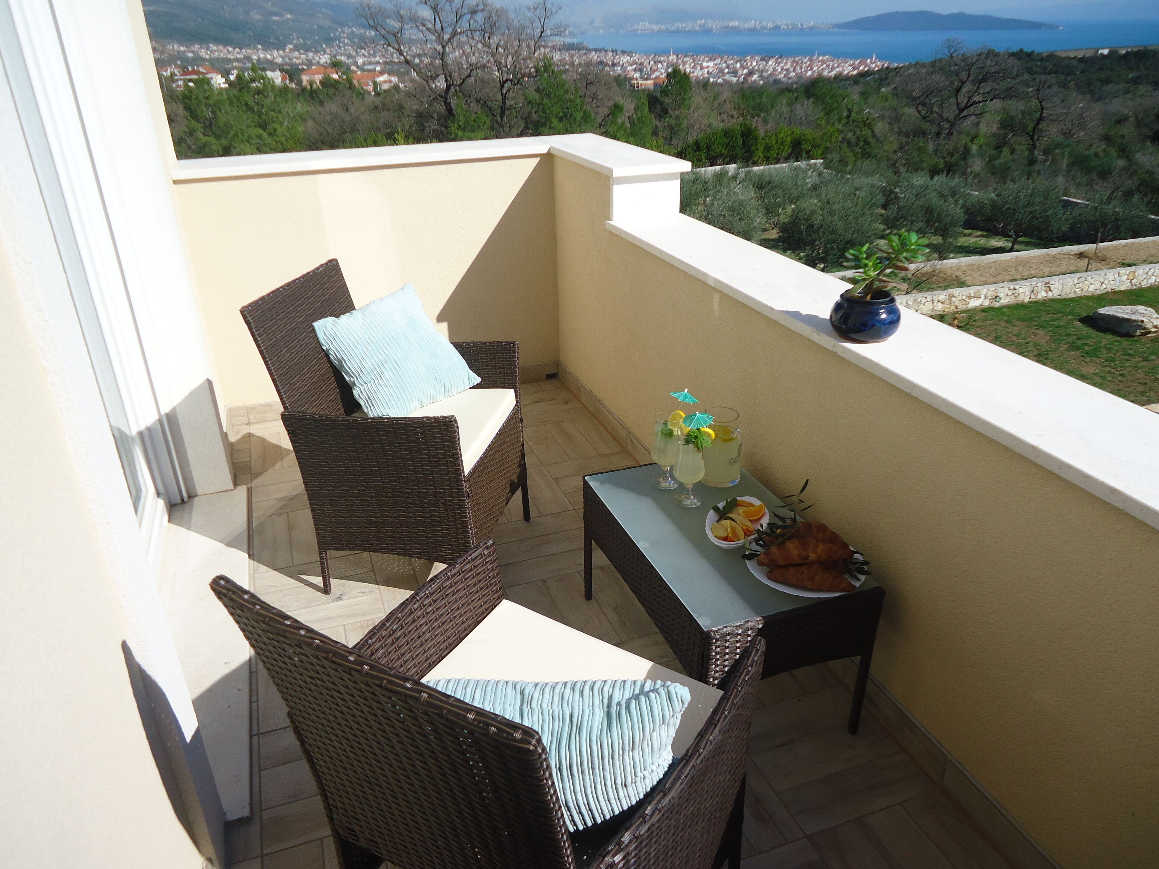 Un balcón con mobiliario de mimbre, una pequeña mesa con bebidas y fruta, y vistas a un pueblo costero.