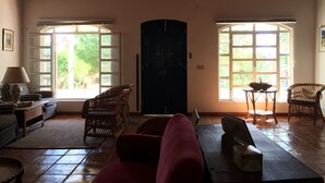Un vaste salon doté d’un haut plafond en bois, de deux grandes fenêtres offrant une vue sur la verdure, d’une porte sombre et de meubles en osier.