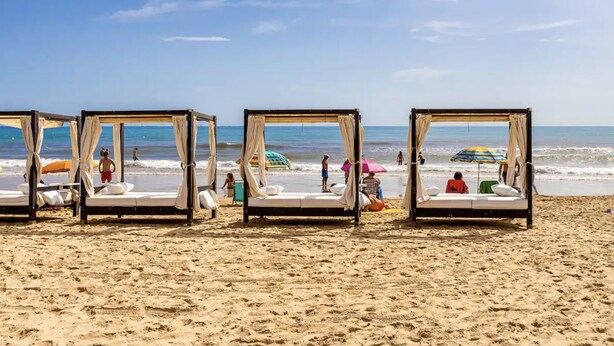 Ein Strand mit vier Holz-Sonnenschirmen, die in einer Reihe angeordnet sind, jeweils mit einer Matratze und Kissen.
