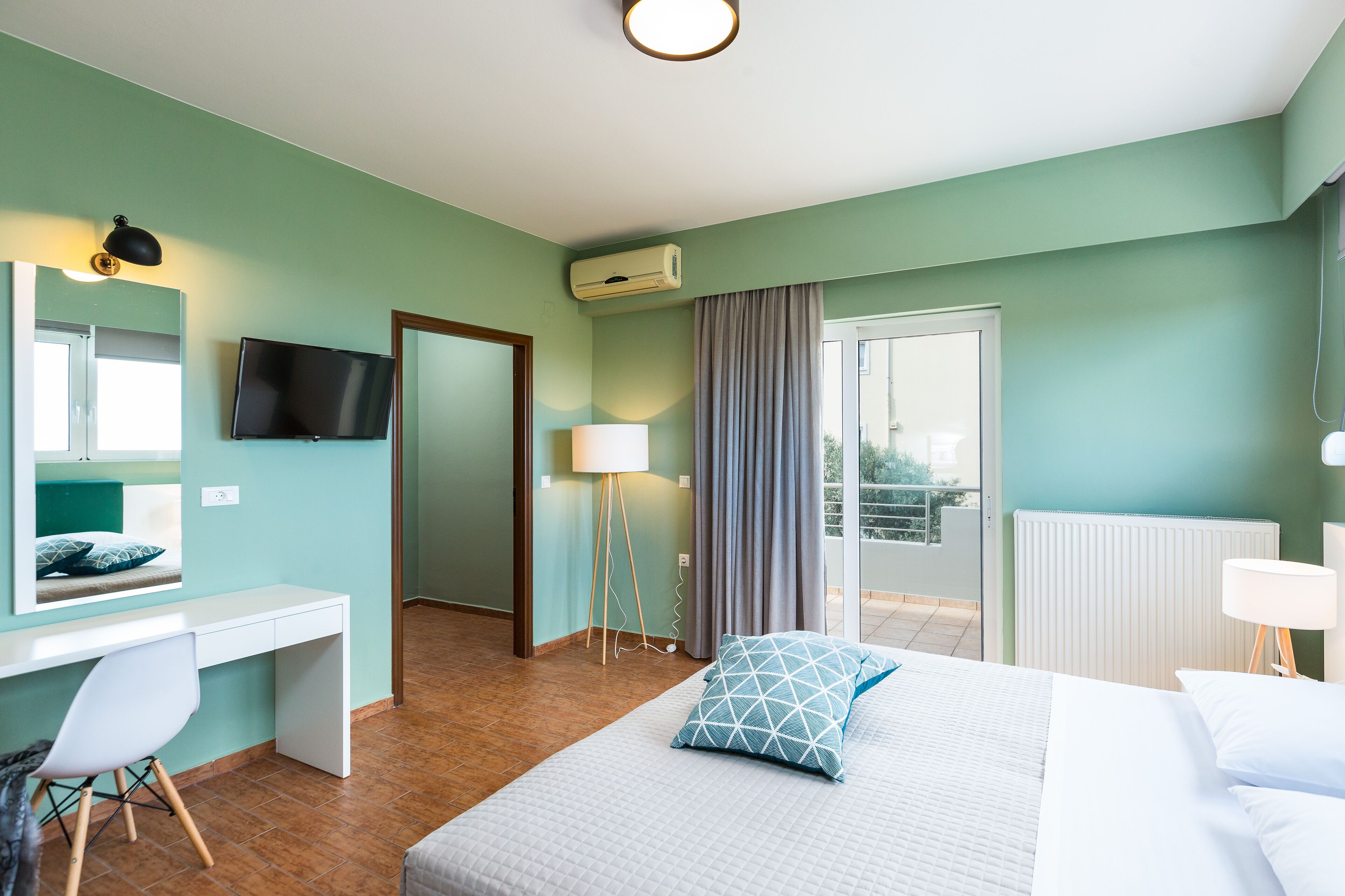 A hotel room with a bed, a desk, a chair, a TV, a lamp, and a view of the outdoors.