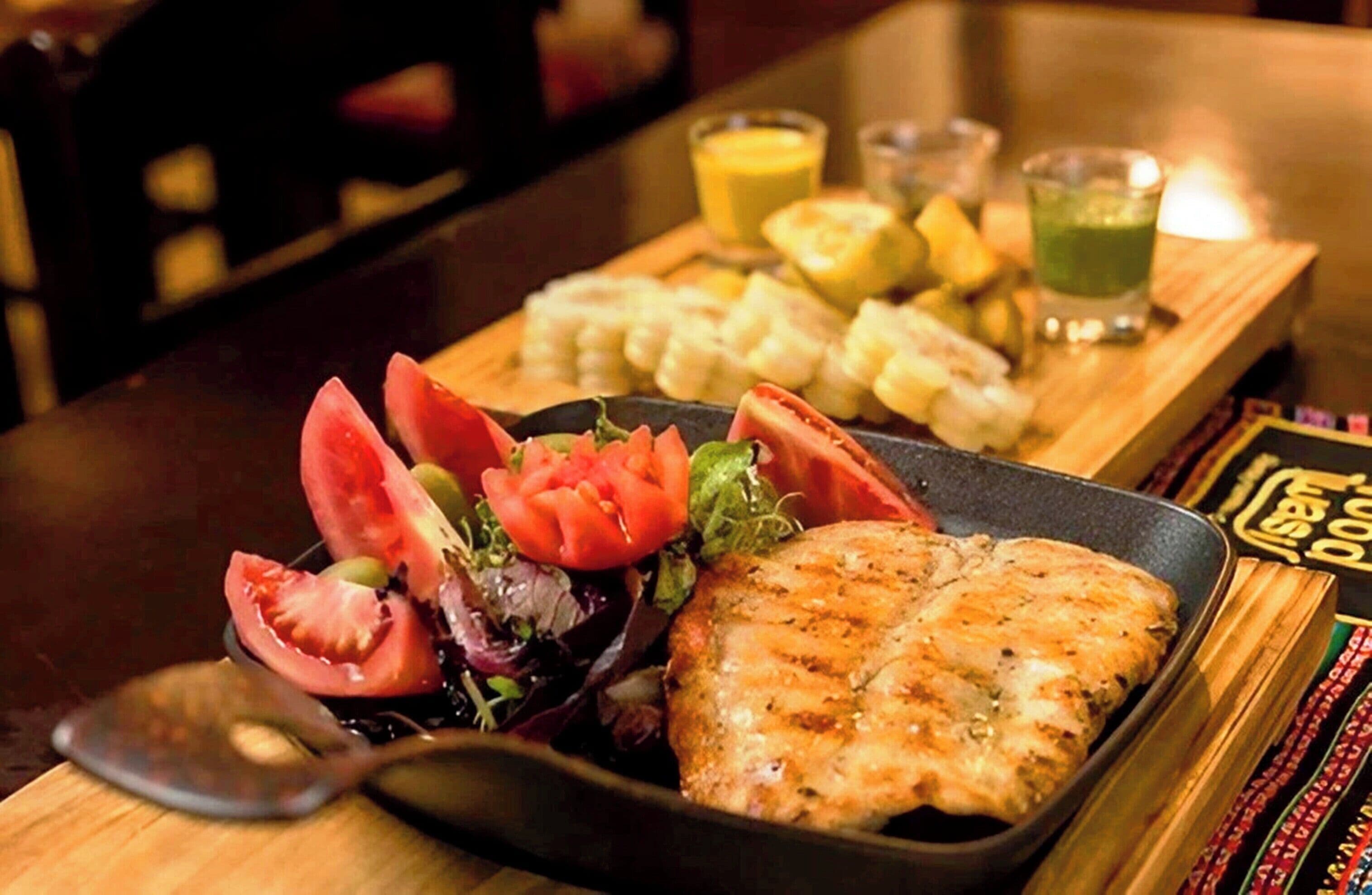 A meal with grilled chicken, tomatoes, and salad on a black plate, accompanied by corn, lemon slices, and a green drink on a wooden board.