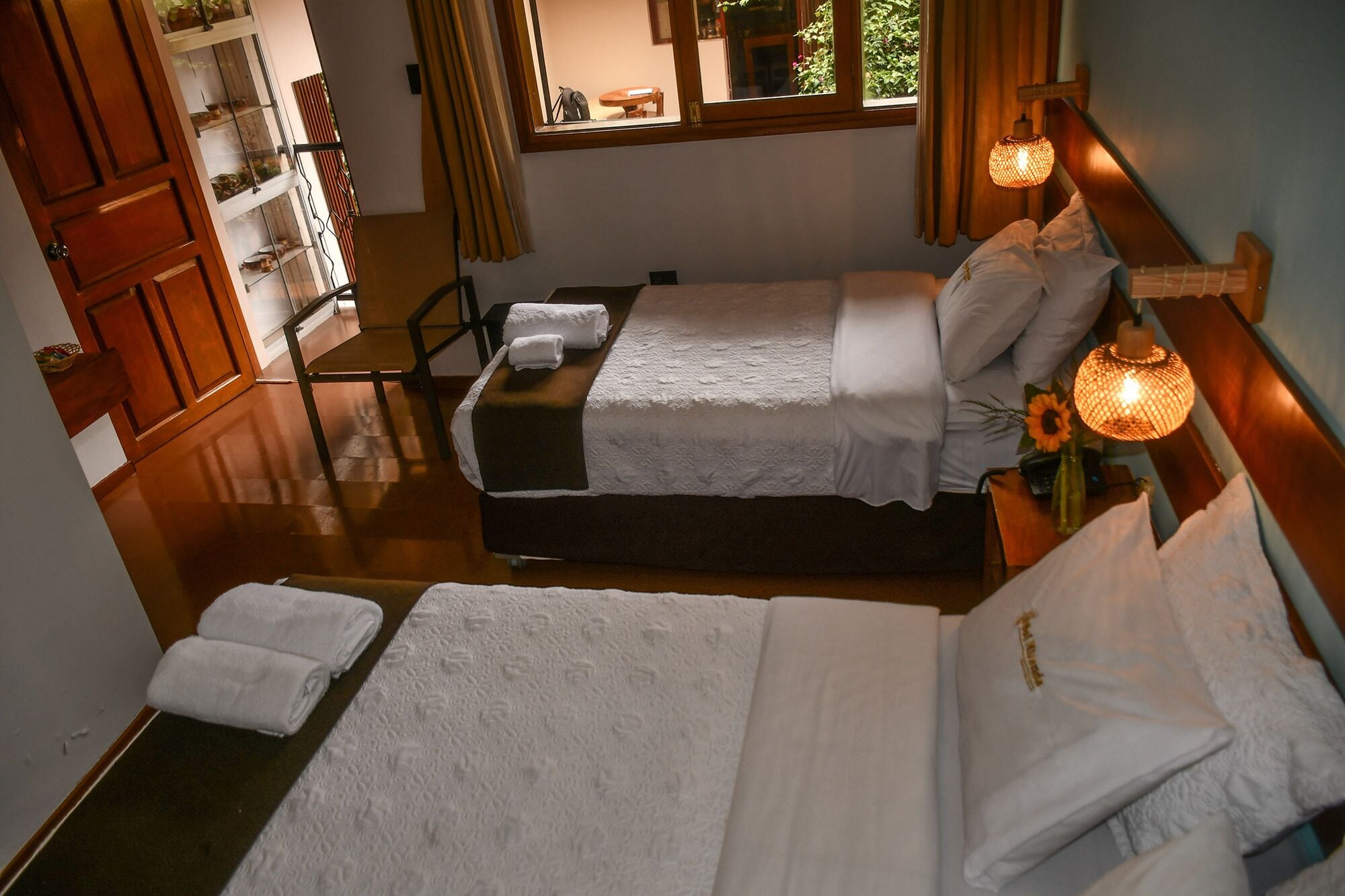 A hotel room with two beds, a wooden floor, a window with curtains, and a bedside lamp.