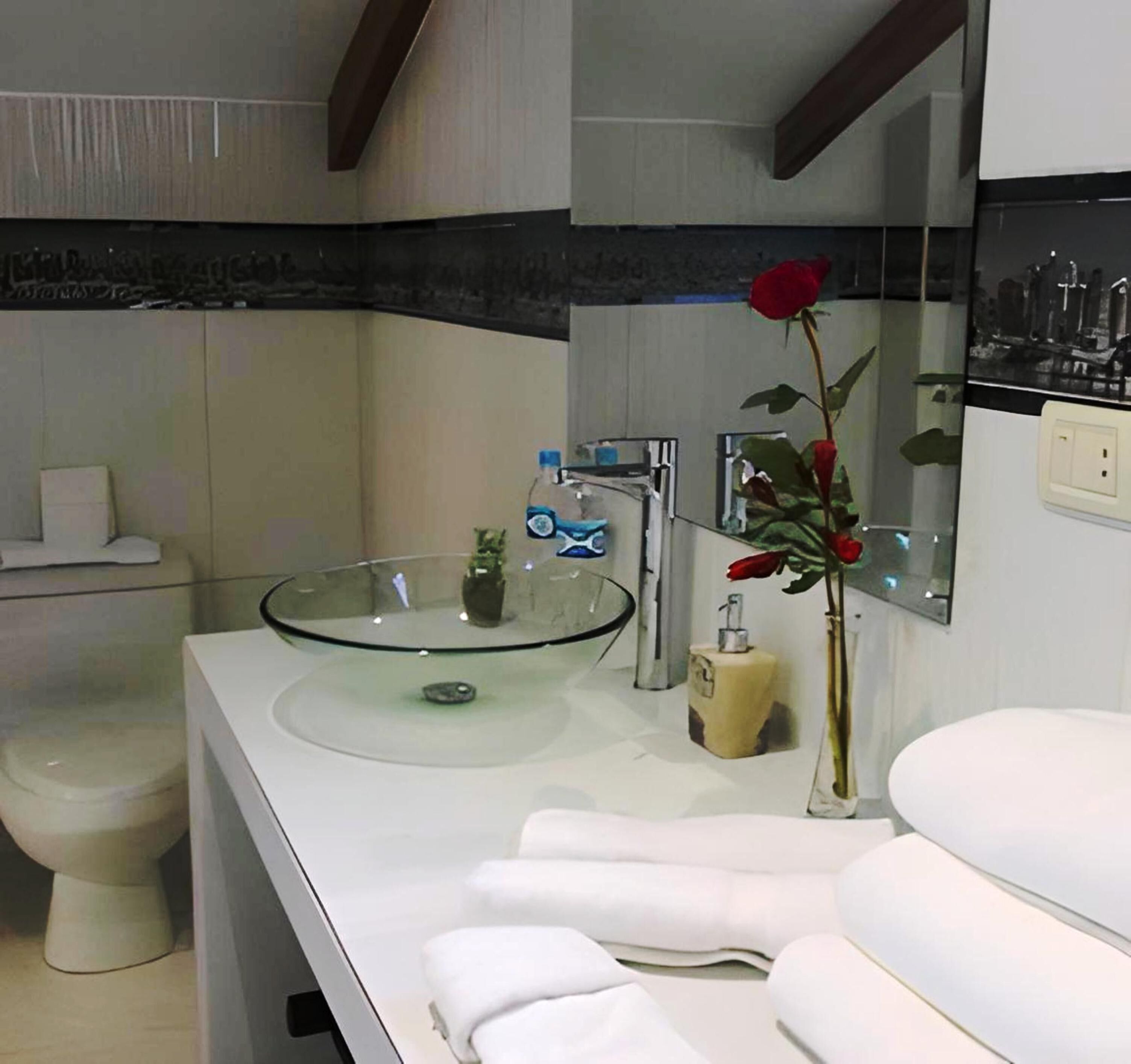 Un baño moderno con un lavabo de vidrio, elementos blancos y una rosa roja en un jarrón.