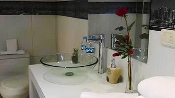 Un baño moderno con un lavabo de vidrio, elementos blancos y una rosa roja en un jarrón.