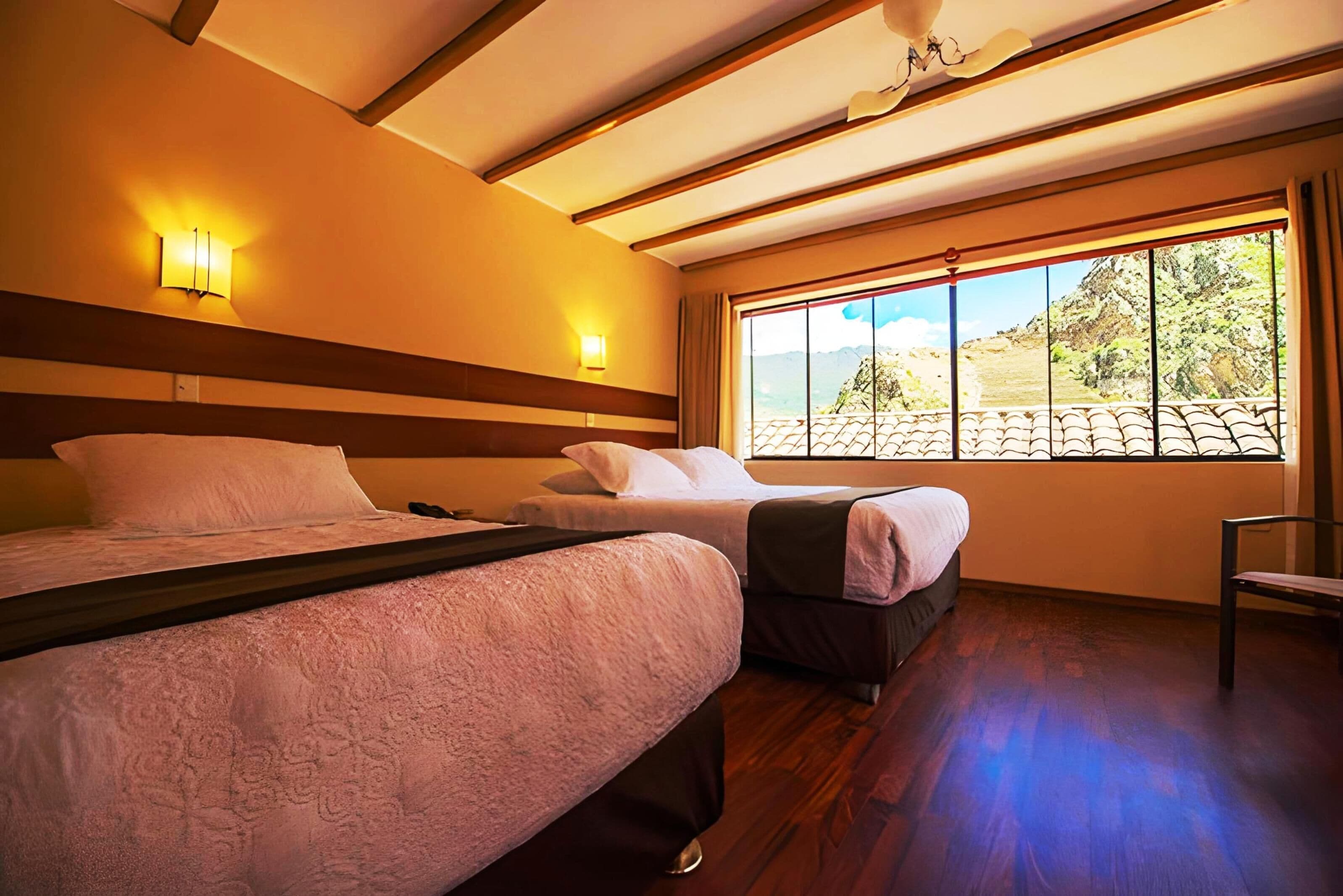 A hotel room with two beds, wooden flooring, a large window with a mountain view, and wall-mounted lights.