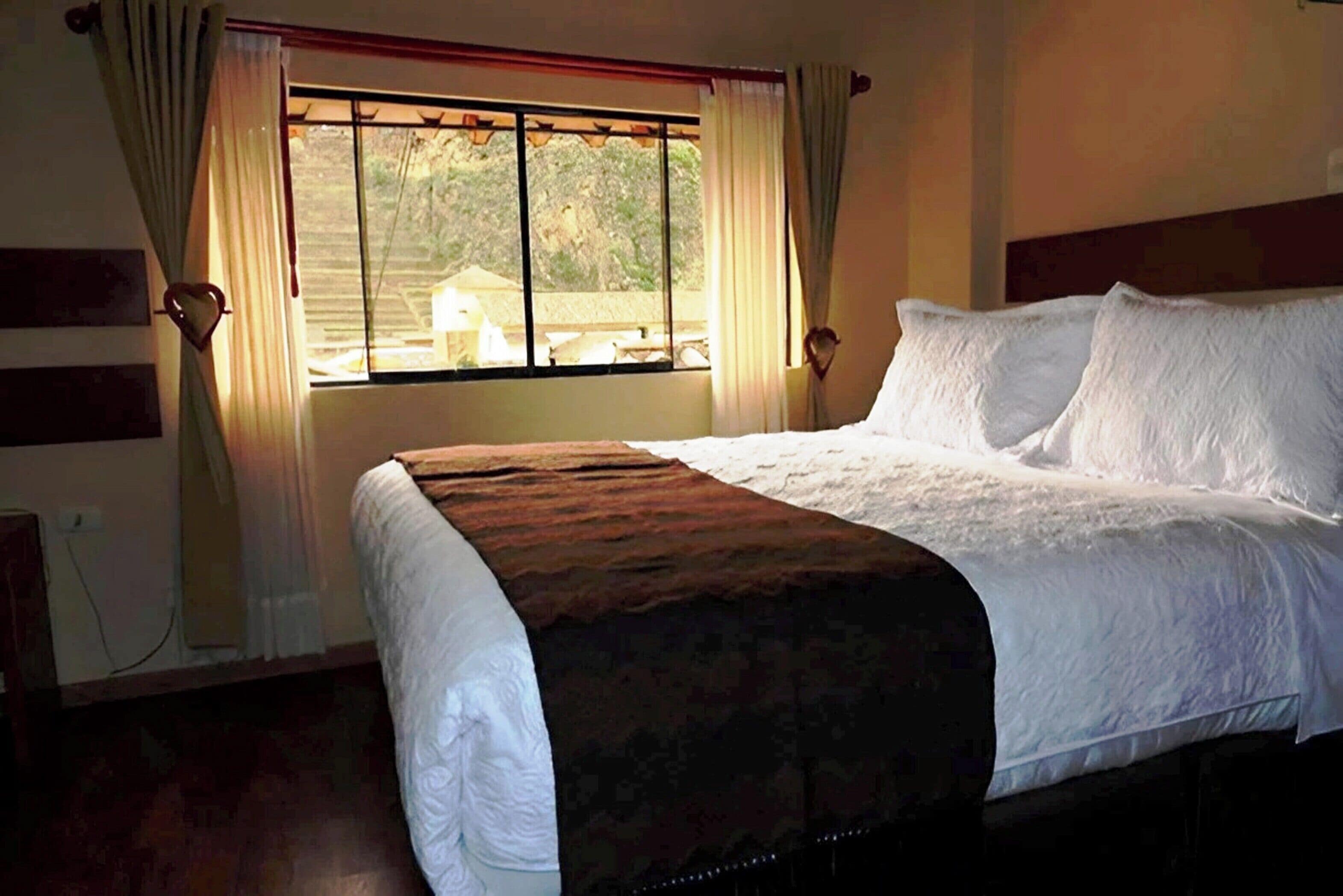 Una cama bien hecha con sábanas blancas y una manta marrón, una ventana con cortinas, y vista a un edificio.