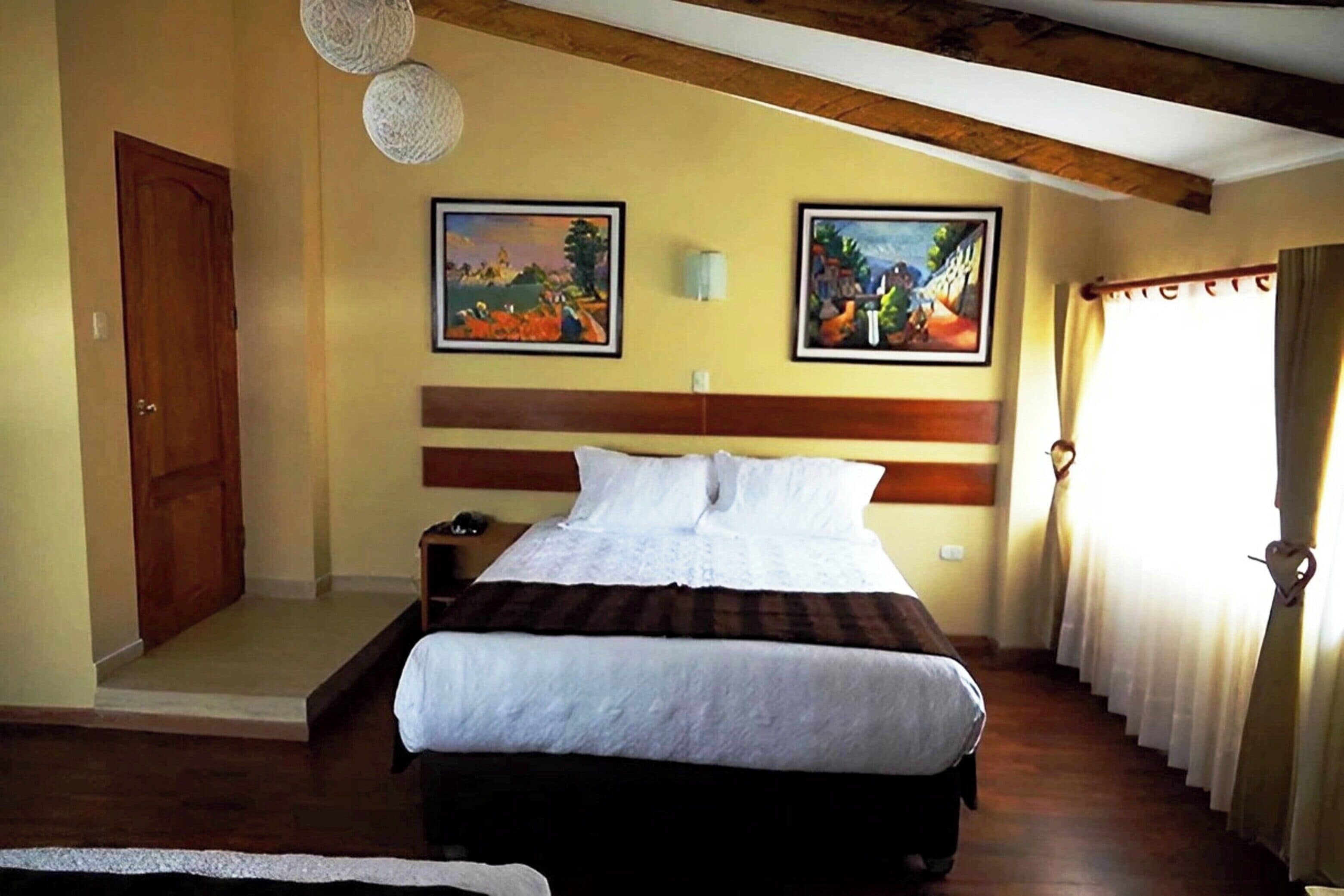 Quarto de hotel com cama, cabeceira de madeira, duas pinturas na parede, mesa de cabeceira e janela com cortinas.