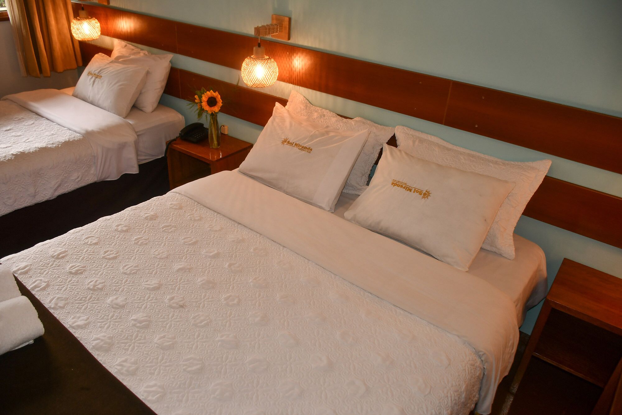 A hotel room with two single beds and one double bed, each with white linens and pillows. There is a nightstand with a lamp and a vase with sunflowers.