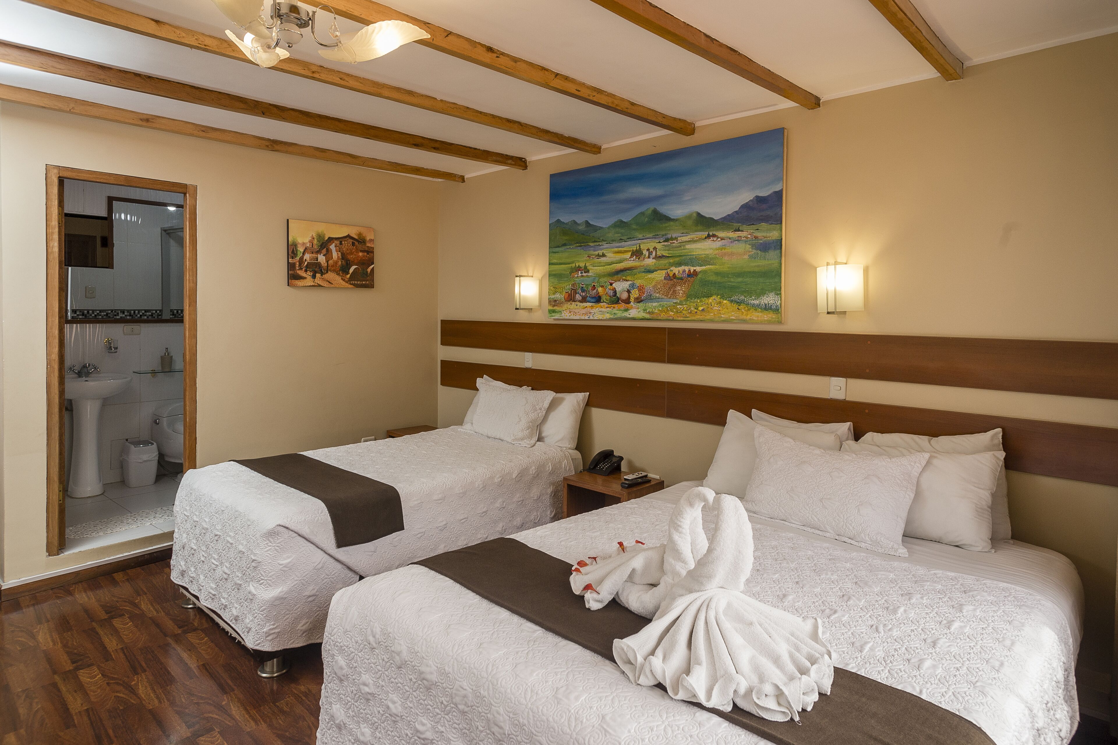 A hotel room with two beds, wooden beams on the ceiling, a painting on the wall, and a bathroom visible through an open door.