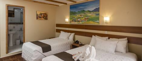 A hotel room with two beds, wooden beams on the ceiling, a painting on the wall, and a bathroom visible through an open door.