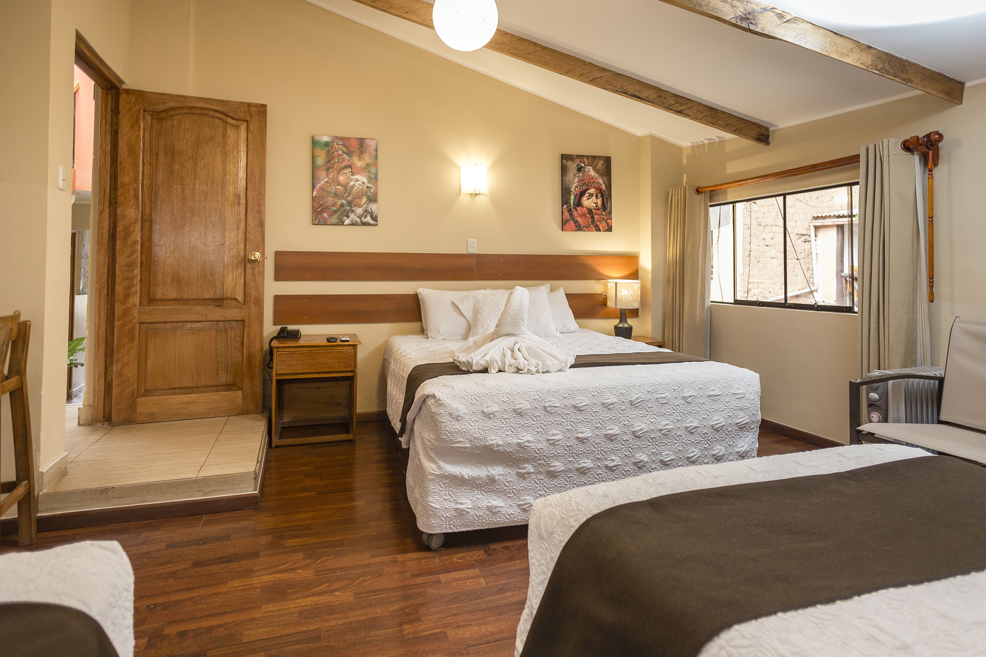 A hotel room with two beds, wooden flooring, a wooden door, a window with curtains, and wall-mounted artwork.