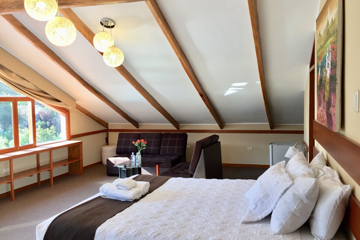 A bedroom with a large bed, a sofa, a table with a vase, and a window with a view of greenery.