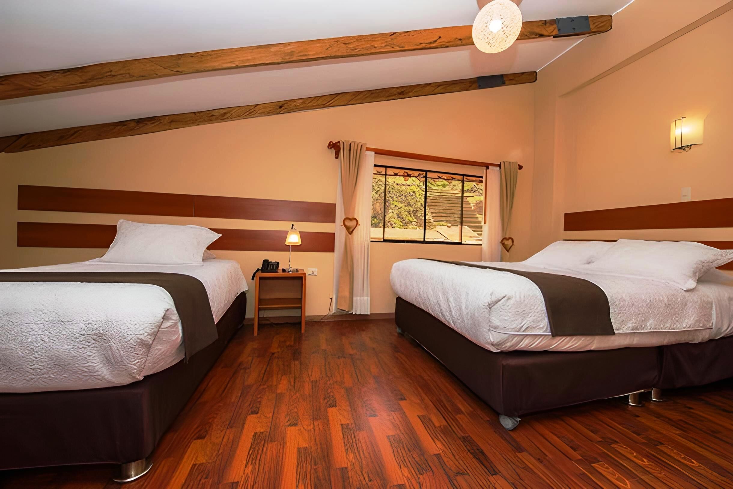 Una habitación con dos camas, suelo de madera, una mesita, una lámpara y una ventana con cortinas.