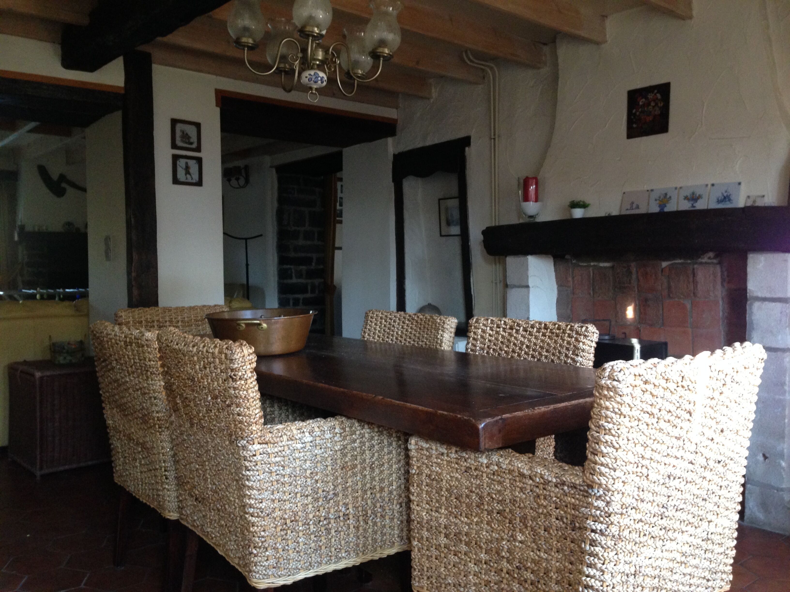 Uma sala de jantar com uma mesa de madeira e cadeiras de fibra trançada, um candelabro e uma lareira de tijolos.