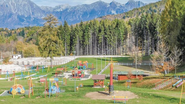 Un parque infantil con diversas estructuras de juego, rodeado de césped y árboles, con montañas al fondo.