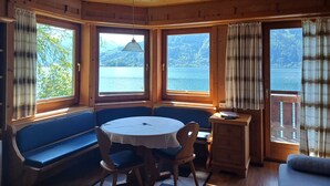 A cabin with a round table and benches, large windows with curtains, and a view of a lake and mountains. - Waterfront Apartment 1 (Zell am See)