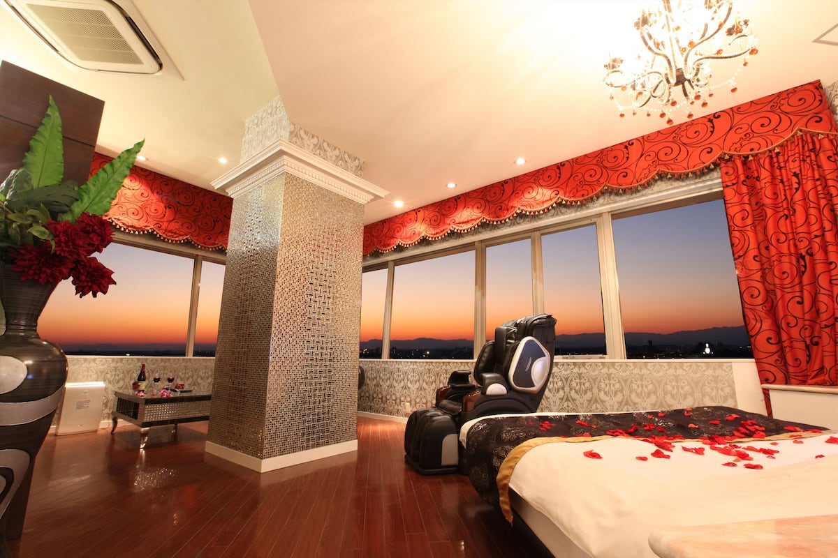 A hotel room with a large bed, a massage chair, a view of the sunset, and a decorative vase with flowers.