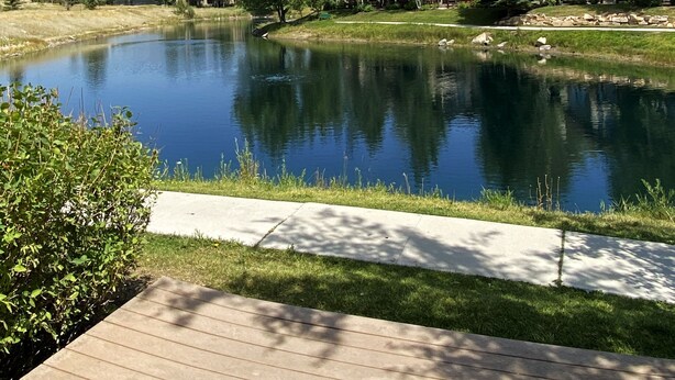 A serene lakeside view with a wooden deck, lush greenery, and modern houses in the background. - 2025 Updates - Modern Townhome In Deer Valley With Shuttle Service (Park City)