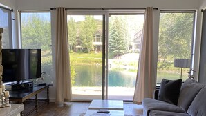 A living room with a sofa, a coffee table, a TV, and a fireplace. There is a large window with curtains, and a view of a pond and trees outside. - 2025 Updates - Modern Townhome In Deer Valley With Shuttle Service (Park City)