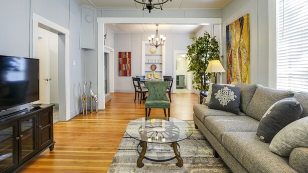 A living room with a television, sofa, and glass coffee table. There is a dining area in the background with a chandelier. - Downtown Historic Charm with Modern Comfort (Prescott)