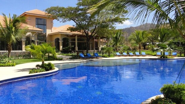 A pool area with a clear blue pool, lounge chairs, and a building with a tiled roof. - Beautiful Penthouse Condo with stunning views that occupies whole floor (Coco)