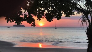 A beach at sunset with calm waters, a boat in the distance, and a palm tree in the foreground. - Beautiful Penthouse Condo with stunning views that occupies whole floor (Coco)