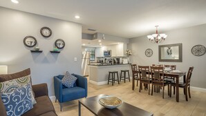 A modern living room with a blue sofa, a dining area with wooden chairs, a kitchen in the background, and decorative wall mirrors.