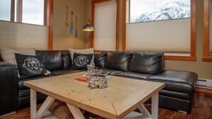 Ein Wohnzimmer mit einer schwarzen Ledersofa, einem Holztisch und Blick auf schneebedeckte Berge durch die Fenster.