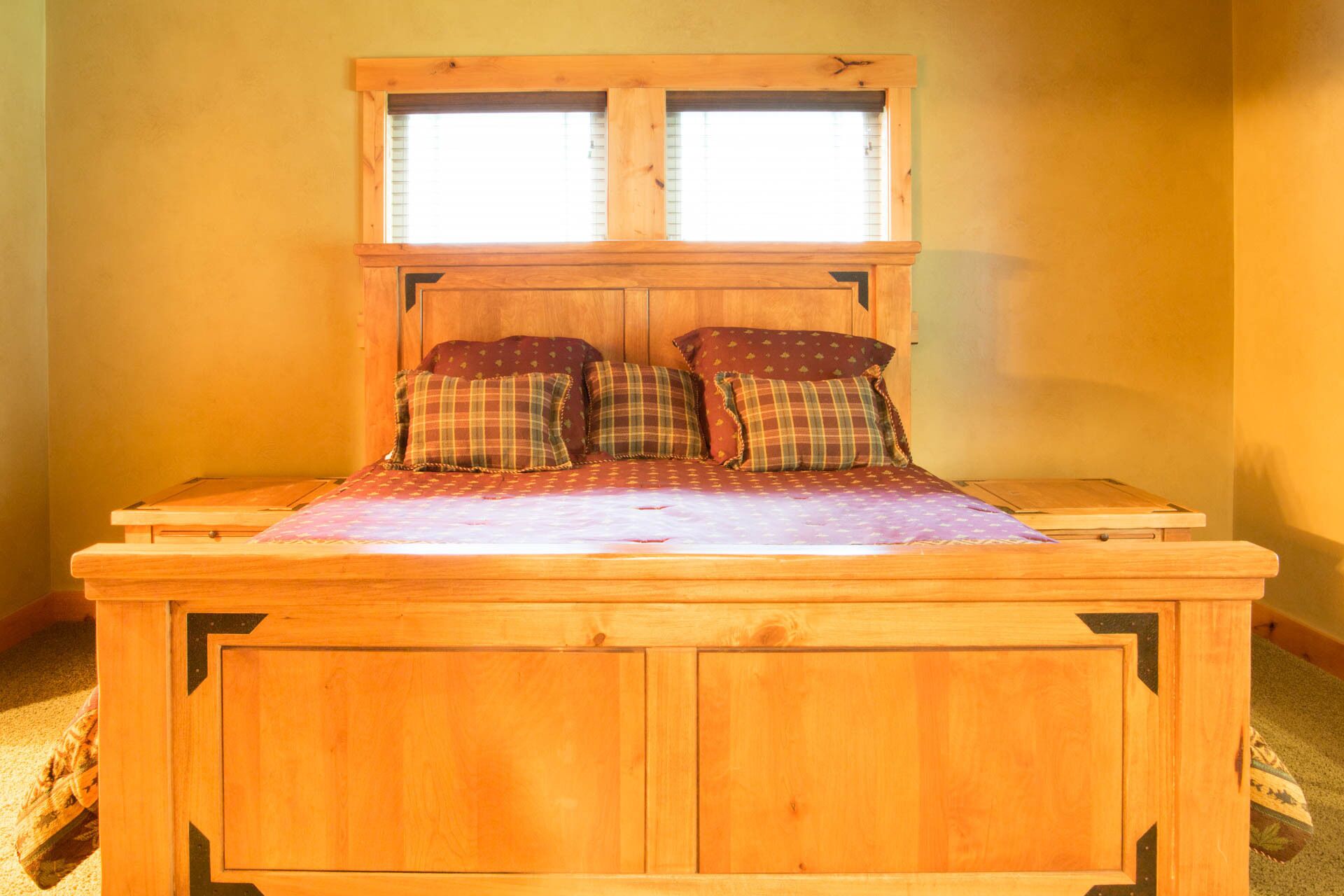 A wooden bed with a headboard, two windows, and plaid bedding.