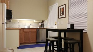 A compact kitchen with wooden cabinets, a dishwasher, a coffee maker, and a small dining table with stools.